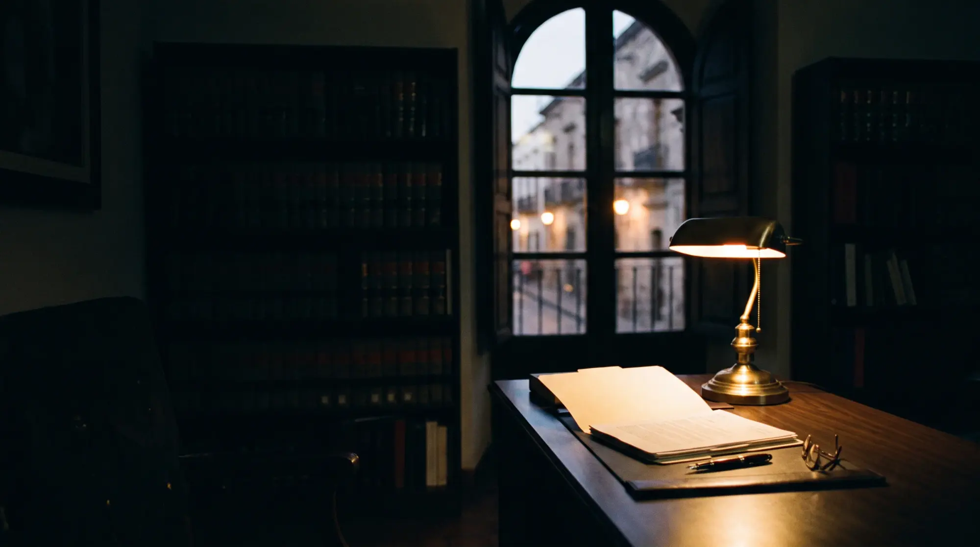 El despacho de noche: una mesa de madera con una carpeta abierta bajo una lámpara de latón, estanterías de libros en penumbra y una ventana a una calle del casco antiguo