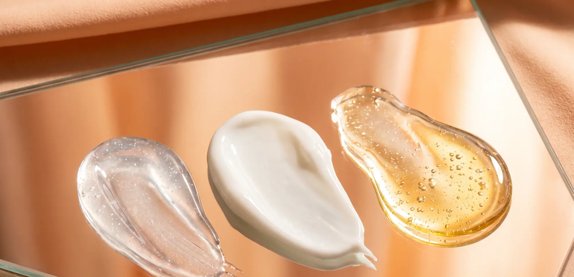 Tres texturas sobre un cristal: un gel transparente, una crema blanca y un aceite dorado